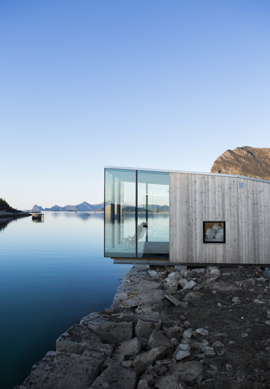 A modern cabin situated right by the water's edge, with views of mountains under a clear sky.